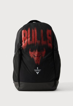 Sac à dos noir des Chicago Bulls avec le nom de l’équipe en rouge et le logo du taureau, des poches latérales en maille, une poignée supérieure et un compartiment avant zippé.