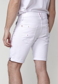 Shorts de mezclilla blanca con puños enrollados, que presentan dos bolsillos traseros, acentos de costura roja y una cinturilla visible con cierre de botón.