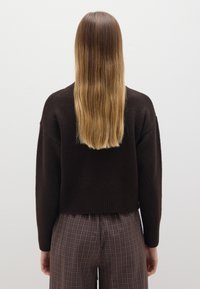 Brauner gestrickter Pullover mit langen Ärmeln und einer verkürzten Silhouette, kombiniert mit braunen karierten Hosen, die an den Knöcheln einen Schlitz haben.
