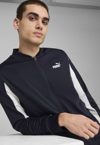 Marineblauwe zip-up trainingsjack met witte zijkanten, gemaakt van gladde stof. Heeft een hoge kraag en een geborduurd Puma-logo op de borst.