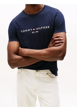 Marineblaue T-Shirt mit Rundhalsausschnitt und weißem „TOMMY HILFIGER EST. 1985“ Logo, kurzen Ärmeln und schlanker Passform, kombiniert mit cremefarbenen Hosen.