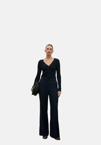 Cardigan aderente blu navy con scollo a V e chiusura con bottoni, abbinato a pantaloni a gamba larga. L'outfit ha una texture morbida, accessoricato con una borsa nera.