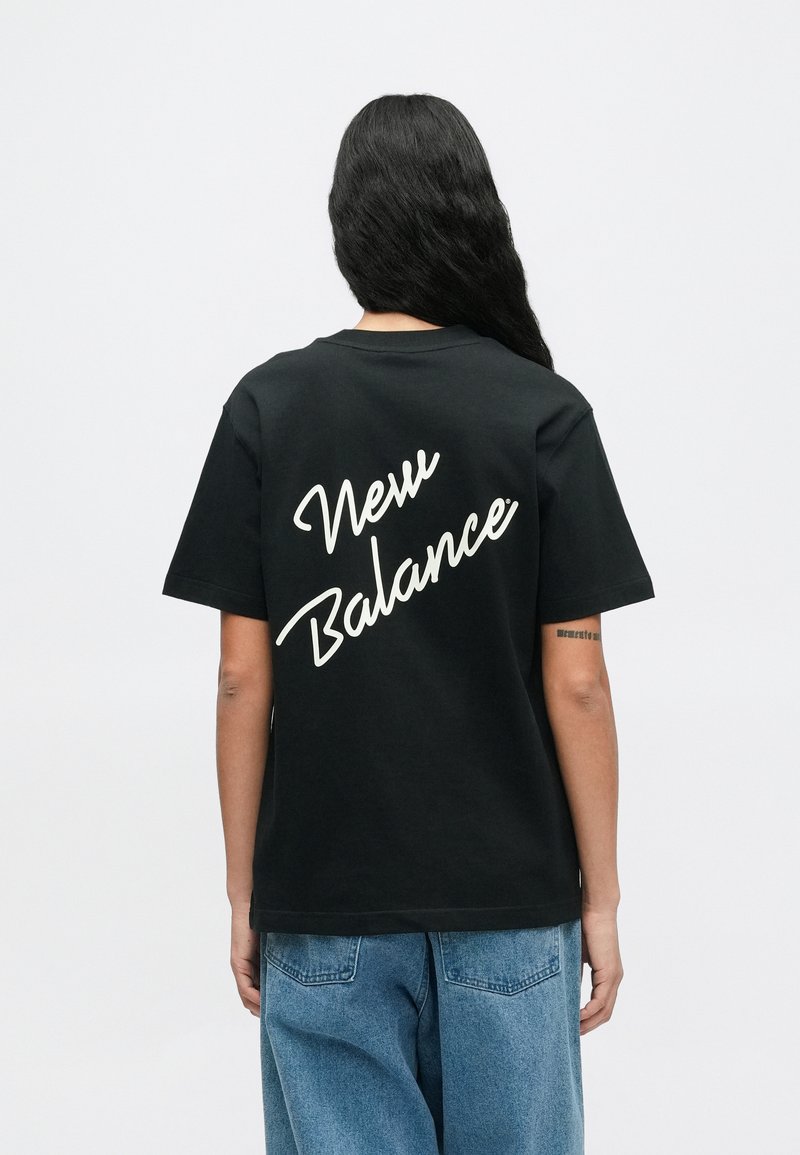 Persona con lunghi capelli scuri che indossa una maglietta nera di New Balance e jeans blu, in piedi di spalle contro uno sfondo semplice.