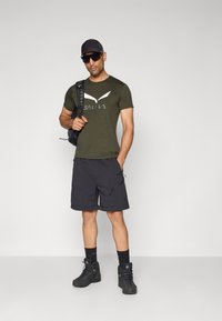Olivengrønn t-skjorte med hvit logo, svarte shorts med glidelås, og svarte tursko. Tilbehør inkluderer en svart ryggsekk og caps.