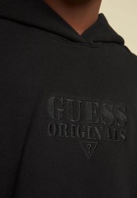 Felpa nera con cappuccio, con il testo ricamato in rilievo "GUESS ORIGINALS" e un piccolo logo triangolare con un punto interrogativo sotto il testo.