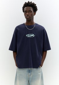 Navy oversized t-shirt made of cotton, featuring a light blue graphic text design "STWD STUDIO," paired with light-wash denim shorts.