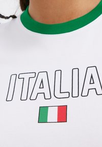 Camiseta blanca con cuello de canalé verde, con la palabra "ITALIA" en letras negras en negrita y un pequeño gráfico de la bandera italiana.