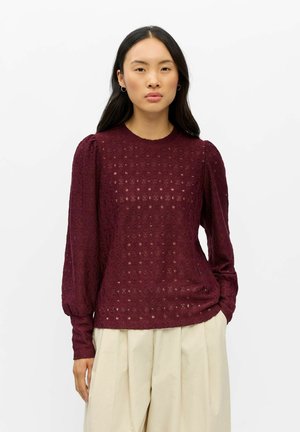 Femme portant un chemisier bordeaux texturé à manches longues avec des épaules bouffantes et un pantalon large beige clair sur un fond uni.