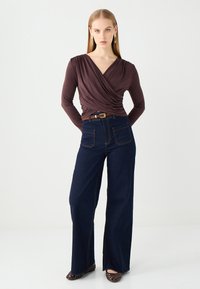 Bruine wikkelblouse met lange mouwen, gecombineerd met hoge, donkere blauwe wijde jeans met voorzakken en contrasterende stiksels.