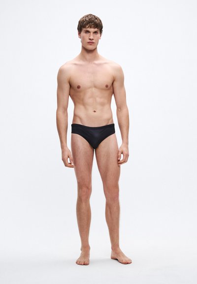 Cuecas de banho pretas com textura suave, design de cintura baixa e cobertura mínima. Modelo de pé descalço contra um fundo simples.