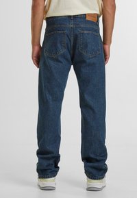 Dark blue denim jeans feature a straight leg design, two back pockets, a leather label on the waistband, and subtle whiskering details.