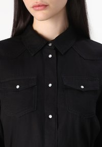Cămașă din denim negru cu guler ascuțit, nasturi cu prindere clară și două buzunare la piept cu detalii din sidef. Țesătură texturată.