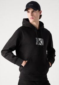Black hooded sweatshirt with a front kangaroo pocket, featuring a silver logo print and drawstring. Cotton material with soft texture.