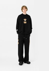 Sudadera negra con un diseño de oso de peluche bordado, combinada con pantalones de chándal negros que presentan detalles similares del oso. Se lleva con zapatos negros texturizados.