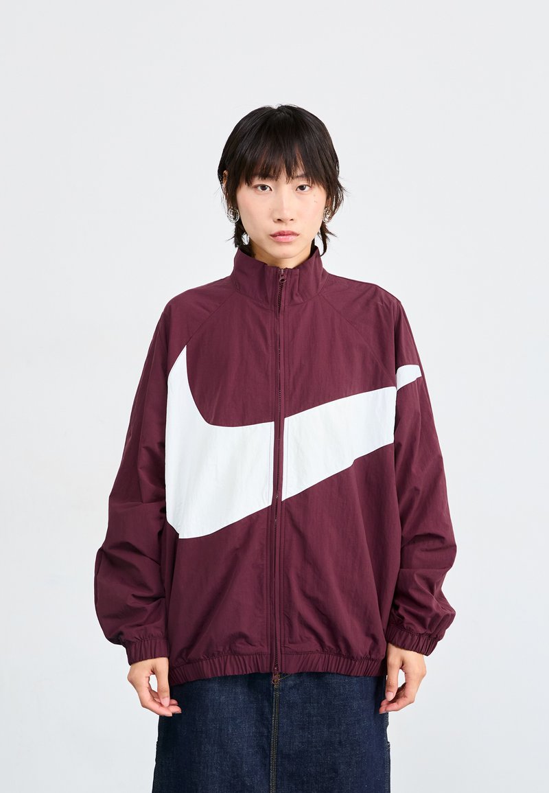 Jachetă maronie cu fermoar și logo alb Nike, având guler înalt, manșete elastice și o croială lejeră. Fabricată din material ușor.