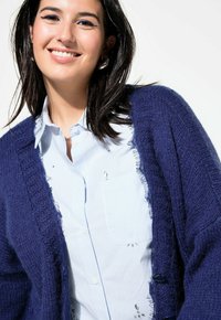 Navy gebreide cardigan met rafelige randen over een lichtblauw gestreept knopshirt. Kenmerkt zich door grote knopen en een gestructureerde stof.