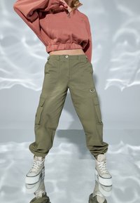 Pantaloni cargo verde oliva con molte tasche e dettaglio a D-ring, abbinati a un maglione rosa corto e scarpe alte beige.