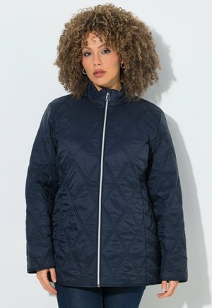 Donna con capelli ricci che indossa una giacca trapuntata blu scuro con una cerniera frontale, in piedi davanti a uno sfondo chiaro.