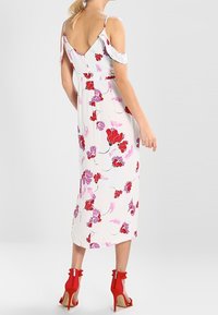 Robe florale blanche avec des fleurs rouges et roses, longueur midi, bretelles dénudées, et sandales à talons rouges. Texture de tissu lisse.