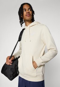 Haut à capuche beige en tissu doux, avec une poche avant, un cordon de serrage pour la capuche et un petit logo blanc ; associé à un sac à épaule noir.