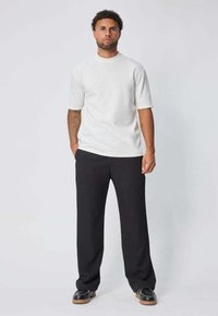 Chemise blanche à manches courtes avec un motif texturé et marbré, associée à un pantalon large noir et des mocassins sombres sur un fond neutre.