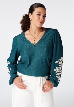 Vrouw met krullend haar die een teal getextureerde top met lange mouwen draagt met beige bloemenborduursel en witte broek, gecompleteerd met gouden sieraden.