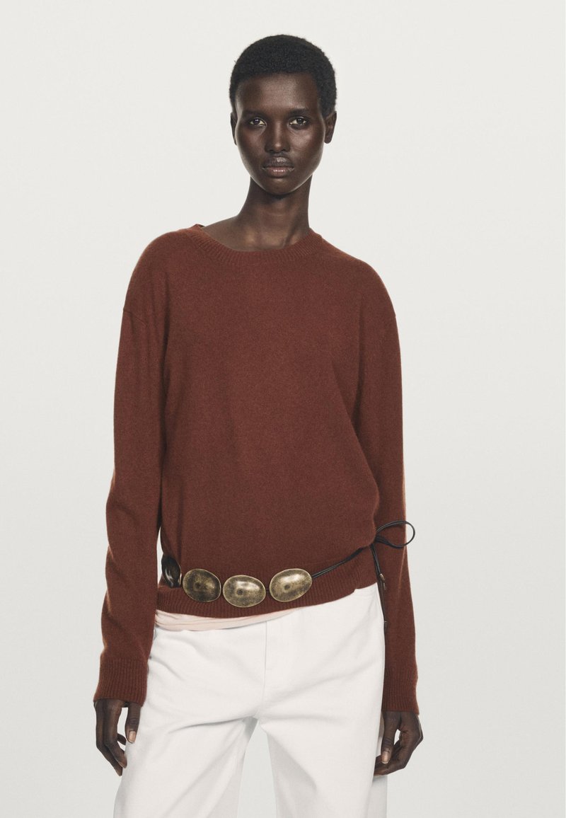 Rostfarbener Pullover mit Rundhalsausschnitt und lockerer Passform, kombiniert mit einem schwarzen Gürtel mit großen metallischen Akzenten. Weiße Hose.