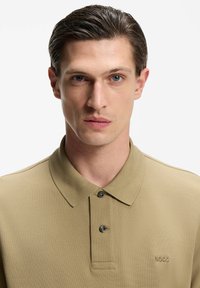 Beżowa koszulka polo o teksturowanym splocie, zapiętym kołnierzykiem i subtelnym logo. Posiada dwa guziki i dopasowany krój.