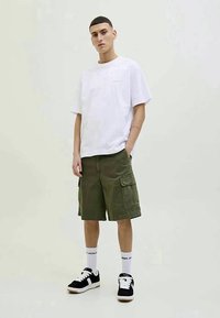 Jovem de pé com as mãos nos bolsos, vestindo uma t-shirt branca oversized, calções de carga verde-oliva, meias brancas e ténis pretos.