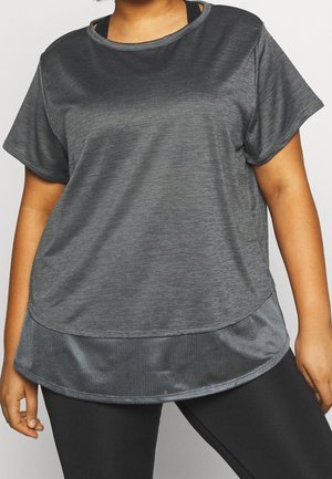 Krągła osoba ubrana w luźną szarą koszulkę z krótkim rękawem z fakturowanej tkaniny i czarne legginsy, stojąca na prostym tle.