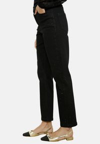 Jeans noirs avec une finition texturée et de petits détails découpés sur le côté, associés à des ballerines noires et dorées avec un design ouvert.