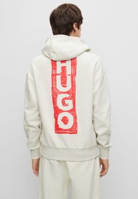 Felpa con cappuccio color crema con stampa testurizzata rossa di "HUGO" sul retro. Presenta una vestibilità comoda e polsini a costine alle maniche.