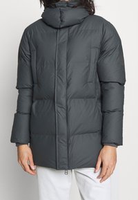 Chaqueta acolchada gris oscuro con cuello alto, paneles cosidos, bolsillos frontales y cierre de cremallera, que presenta una textura suave y mate.
