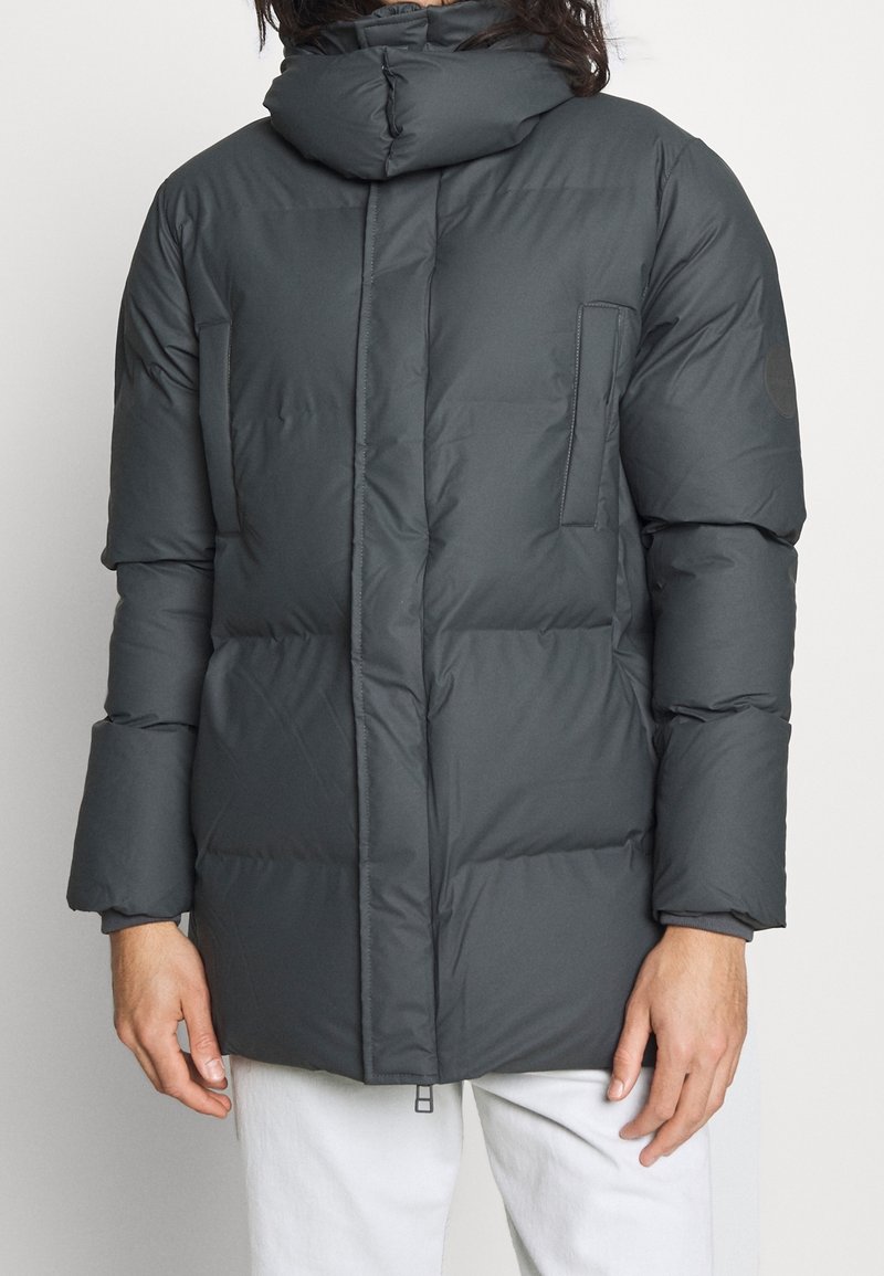 Chaqueta acolchada gris oscuro con cuello alto, paneles cosidos, bolsillos frontales y cierre de cremallera, que presenta una textura suave y mate.