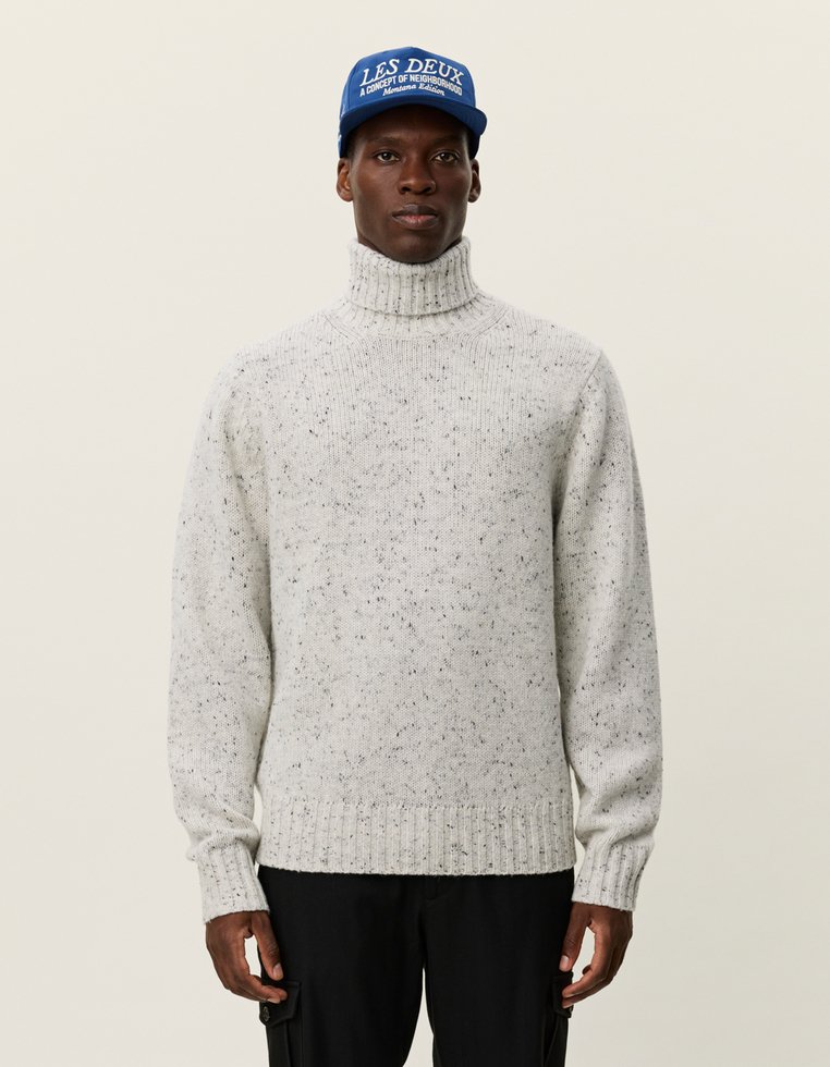 Hellgrauer, gestrickter Rollkragenpullover mit geripptem Saum, der dunkle Flecken aufweist. Kombiniert mit einer blauen Mütze, die weißen Text zeigt.