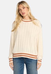 Pull rayé crème avec un tissu texturé, présentant un ourlet et des poignets marron en côtes, et un design à coupe décontractée. Associé à un jean bleu.
