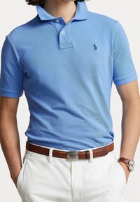 Jasnoniebieska koszulka polo z krótkim rękawem, z teksturowanej tkaniny, z kołnierzykiem na dwa guziki oraz małym haftowanym logo po lewej stronie klatki piersiowej. W zestawieniu z białymi spodniami i brązowym paskiem.