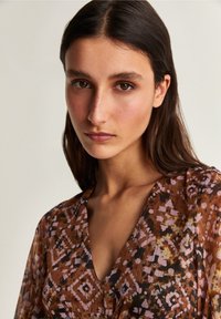 Bruine blouse met patroon en V-hals, met paarse en zwarte geometrische ontwerpen. Lichtgewicht, textiel met een vloeiende silhouette.