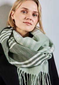 Écharpe vert clair avec des rayures blanches, bords frangés, en texture douce, drapée autour du cou contre un fond de manteau noir.