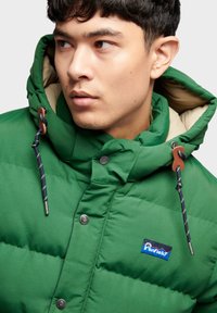 Green puffer jacket with a contrasting beige hood, featuring metal snap buttons and a branded patch on the chest. Textured, insulated fabric.