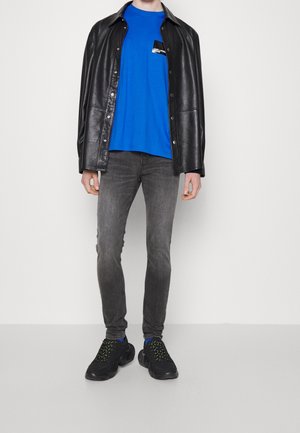 Person trägt eine schwarze Lederjacke über einem blauen T-Shirt, graue Slim-Fit-Jeans und schwarze klobige Sneaker vor einem schlichten weißen Hintergrund.