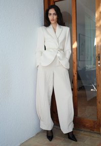 Beige skräddarsydd kavaj med bälte i midjan, i kombination med vida krämfärgade byxor. Modellen står mot en vägg med en bakgrund av en träport.