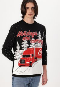 Pletený sveter s čiernym telom, bielymi snehovými vločkami, červeným textom "Vianoce prichádzajú" a červeným nákladiakom Coca-Cola medzi bielymi stromami.