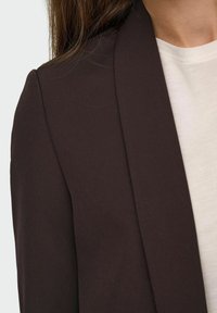Tamno smeđi blazer s glatkom teksturom, s pravilno oblikovanim reverom i zatvaranjem na jedan gumb, nosi se preko svijetle košulje.