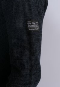 Donkerblauwe gebreide sweater met een gestructureerde afwerking en een zichtbaar zwart stoflabel op de linkermouw, met witte tekst en merkinformatie.