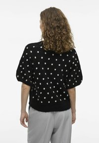 Melnā džemperis ar baltām punktiņām, īsām pūkainām piedurknēm un ribinātu apakšmalu. Mīksta tekstūra. Valkāts ar gaiši pelēkiem biksēm.