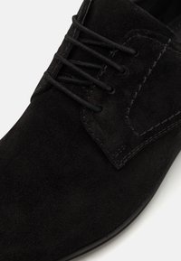 Chaussure en suede noire à lacets avec une texture lisse et un accent en onglet cousu. Présente un design épuré et minimal ainsi qu'une semelle en caoutchouc durable.