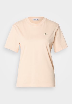 Broskyňovo bavlnené tričko s okrúhlym výstrihom, krátkymi rukávmi a malým zeleným vyšitým logom krokodíla na ľavej strane hrude.