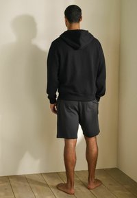 Sudadera con capucha negra y puños acanalados, combinada con pantalones cortos gris oscuro. El modelo está de pie sobre un suelo de madera, descalzo, de frente a una pared sencilla.