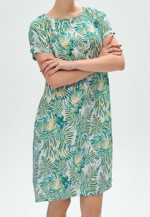 Žena se založenýma rukama, oblečená v krátkém šatě ke kolenům s motivem zelených a modrých tropických listů na bílém pozadí.